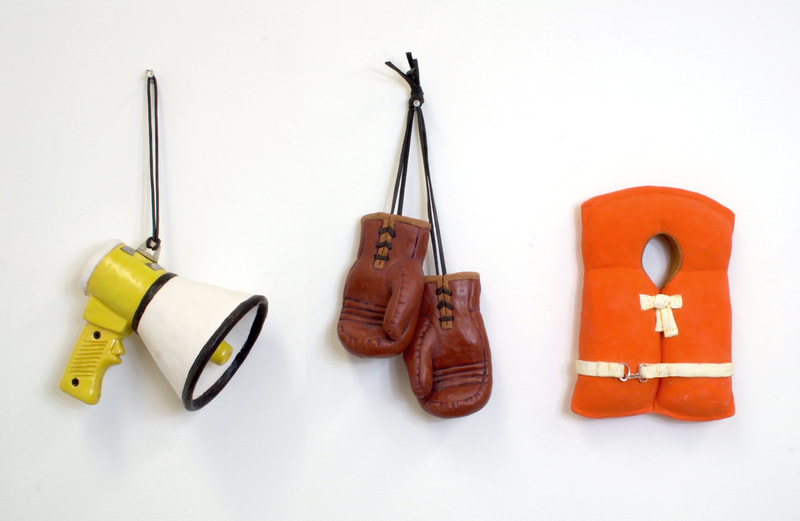 tools for survival, a clay megaphone, boxing gloves and life vest all hanging on a white wall
